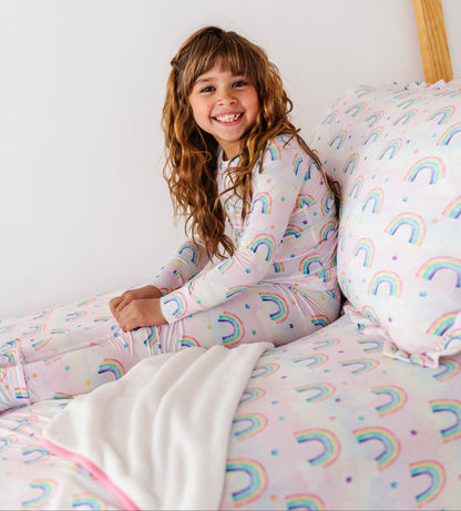 Young girl in rainbow-patterned bamboo pajamas sitting on a bed with matching bamboo bedding.