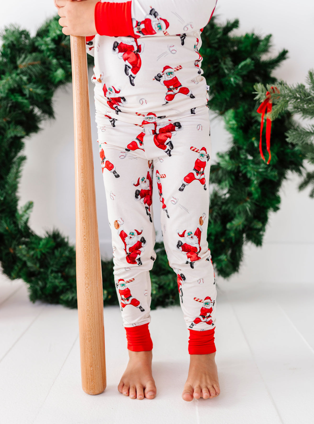 A child wearing a white Christmas pajama set with a Santa Claus print and red cuffs, standing barefoot and holding a wooden bat in front of a green holiday wreath.
