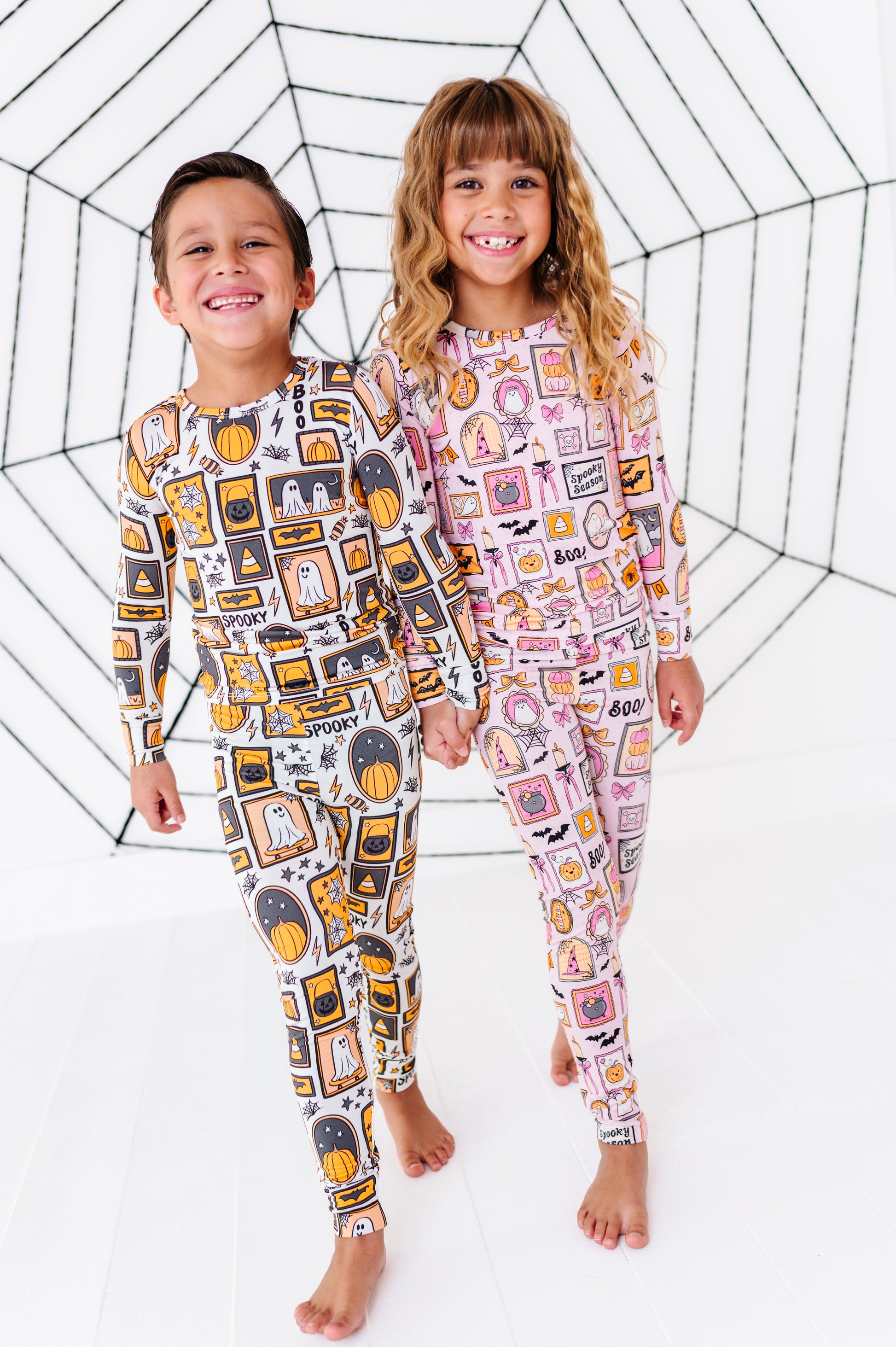 Two children standing barefoot in front of a white studio backdrop with a black spiderweb graphic. The child on the left wears a long-sleeve one-piece pajama with a black, yellow, and gray graphic print, and the child on the right wears a matching long-sleeve one-piece pajama with a pink, white, and orange graphic print.