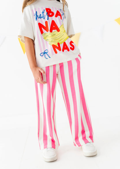 A child wearing a white graphic T-shirt with colorful text and a banana illustration, paired with pink and white vertically striped pants and white sneakers, photographed against a clean white studio background.