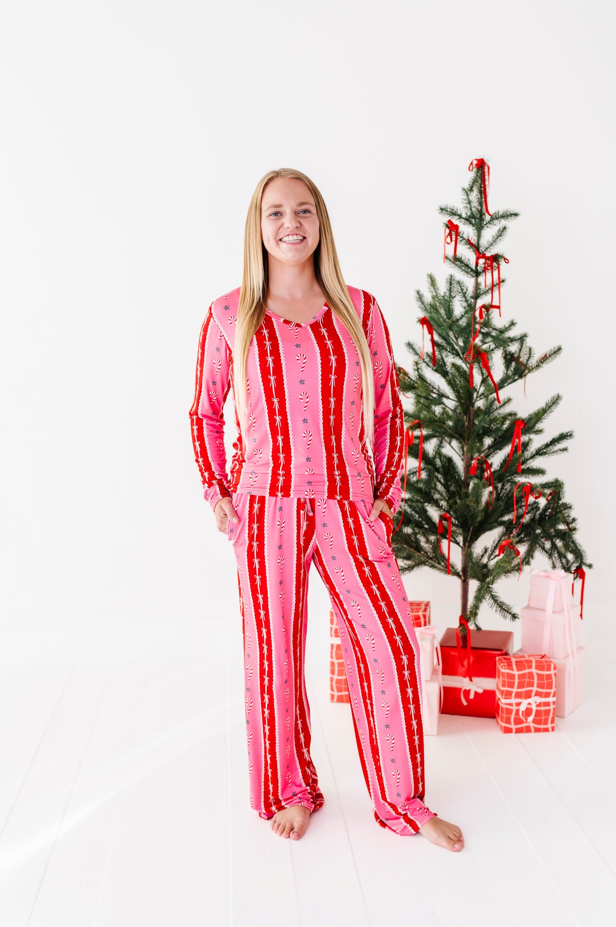 A woman stands barefoot in a bright white room wearing a pink and red striped bamboo pajama set. A decorated Christmas tree with red ribbons and wrapped gifts is visible behind her.