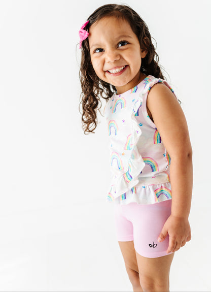 Young girl wearing a sleeveless bamboo cotton peplum top with watercolor rainbow patterns and pink bamboo biker shorts on a white background