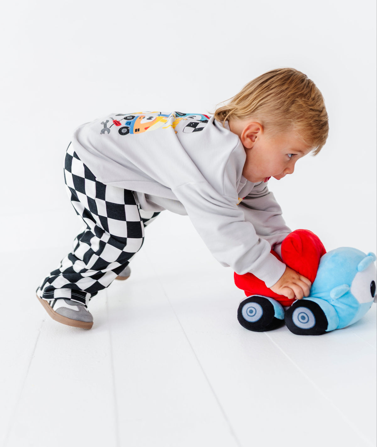 A toddler wearing a light gray long-sleeve shirt and black-and-white checkered pants crawls on a white floor while pushing a red and blue plush toy car.