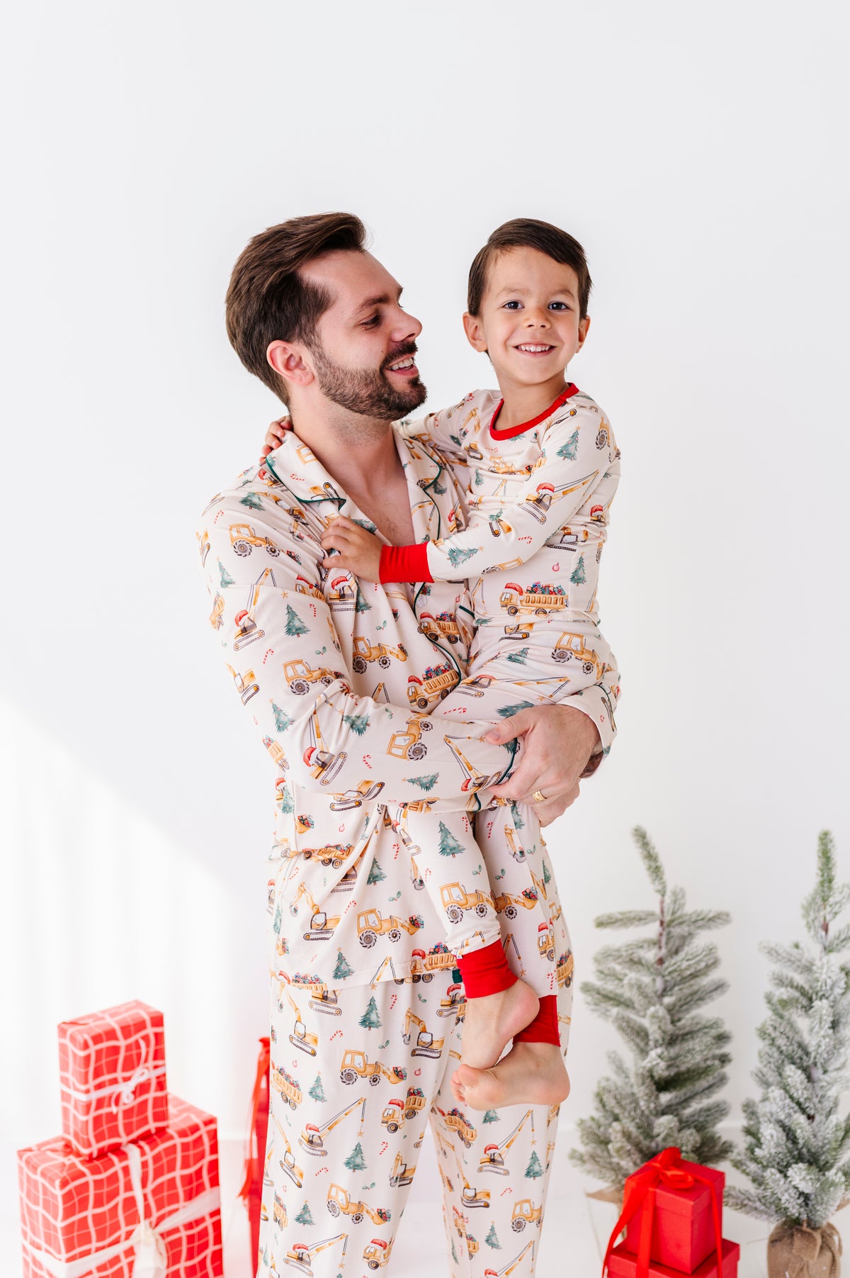 Father and son in matching Christmas construction-themed pajamas standing in front of a Christmas tree and a pile of presents 
