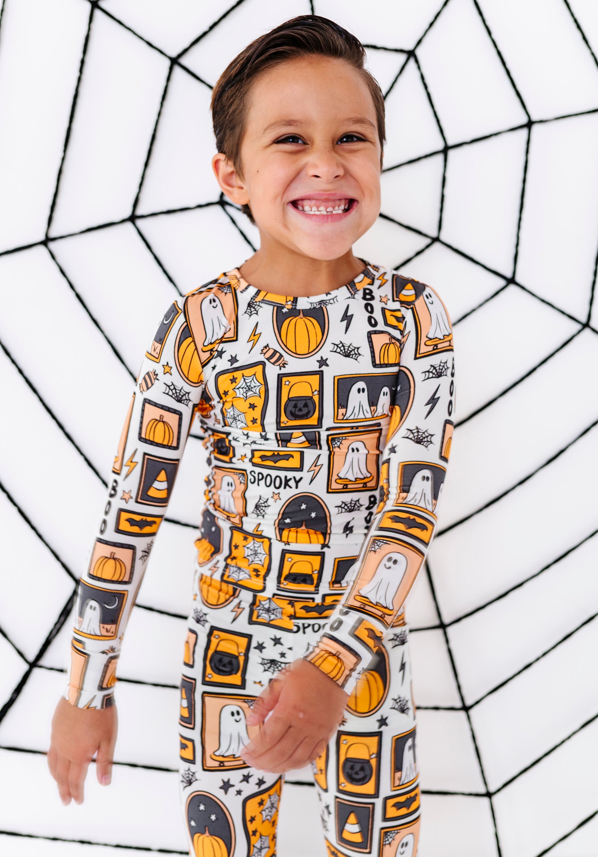 A smiling young boy wearing a long-sleeve Halloween-themed pajama set with an orange, black, and white spooky print, standing in front of a white background with a black spiderweb pattern.