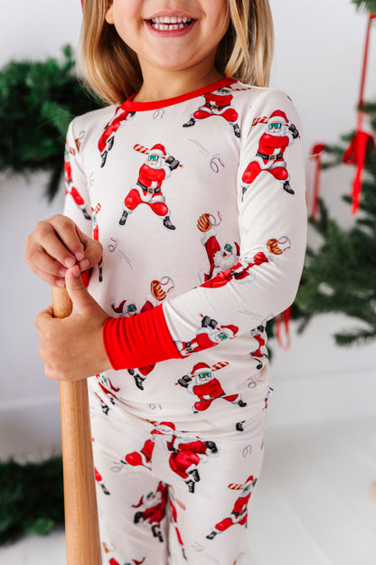 A child wearing a long-sleeve bamboo pajama top and matching pants with an all-over Santa Claus baseball print, holding a wooden bat in a festive indoor setting with Christmas decorations in the background.