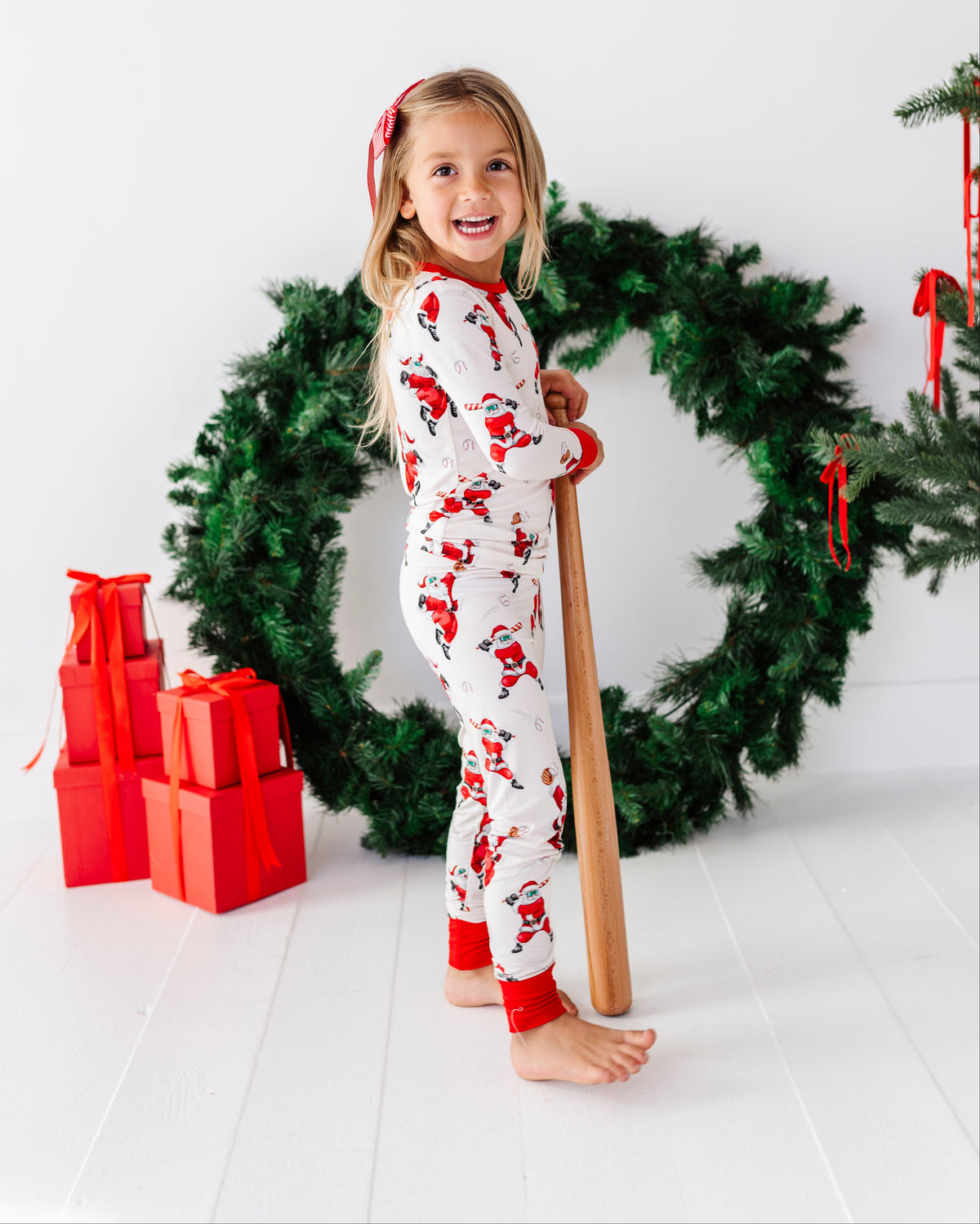 A young girl stands barefoot in a holiday-themed studio setting, wearing a white Christmas pajama set with red Santa Claus prints. She is holding a wooden baseball bat in front of a large green Christmas wreath with red ribbons, with red wrapped gift boxes on the left against a clean white background.