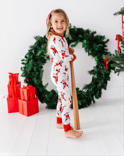 A young girl stands barefoot in a holiday-themed studio setting, wearing a white Christmas pajama set with red Santa Claus prints. She is holding a wooden baseball bat in front of a large green Christmas wreath with red ribbons, with red wrapped gift boxes on the left against a clean white background.