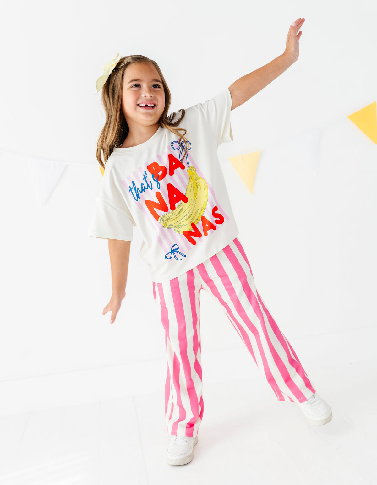 A young girl modeling a white graphic t-shirt that reads 'that's BA NANA'S' with a banana illustration, paired with pink and white vertically striped pants, against a bright white studio background.