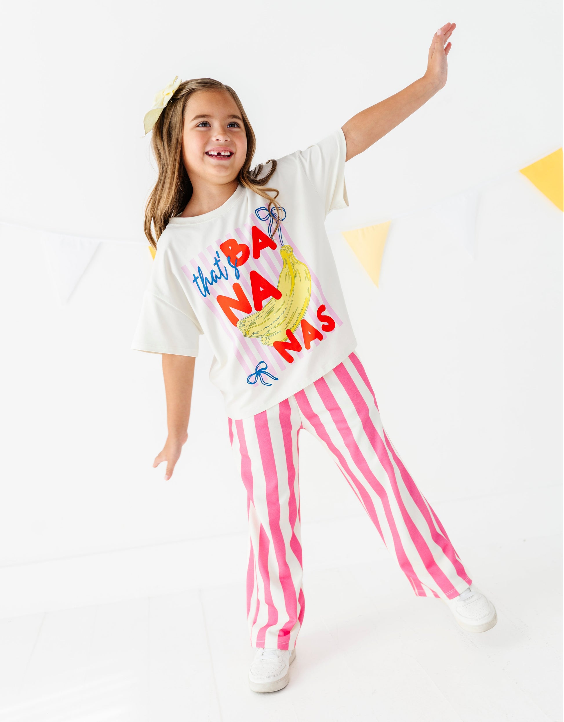 A young girl modeling a white graphic t-shirt that reads 'that's BA NANA'S' with a banana illustration, paired with pink and white vertically striped pants, against a bright white studio background.