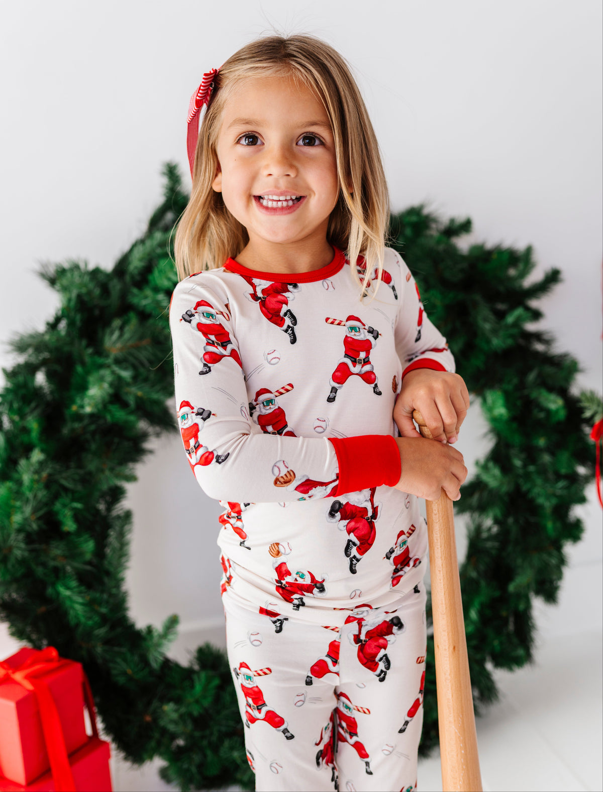 A young girl smiling while wearing a Santa Claus patterned long-sleeve pajama set, holding a wooden stick in front of a green wreath and Christmas tree.