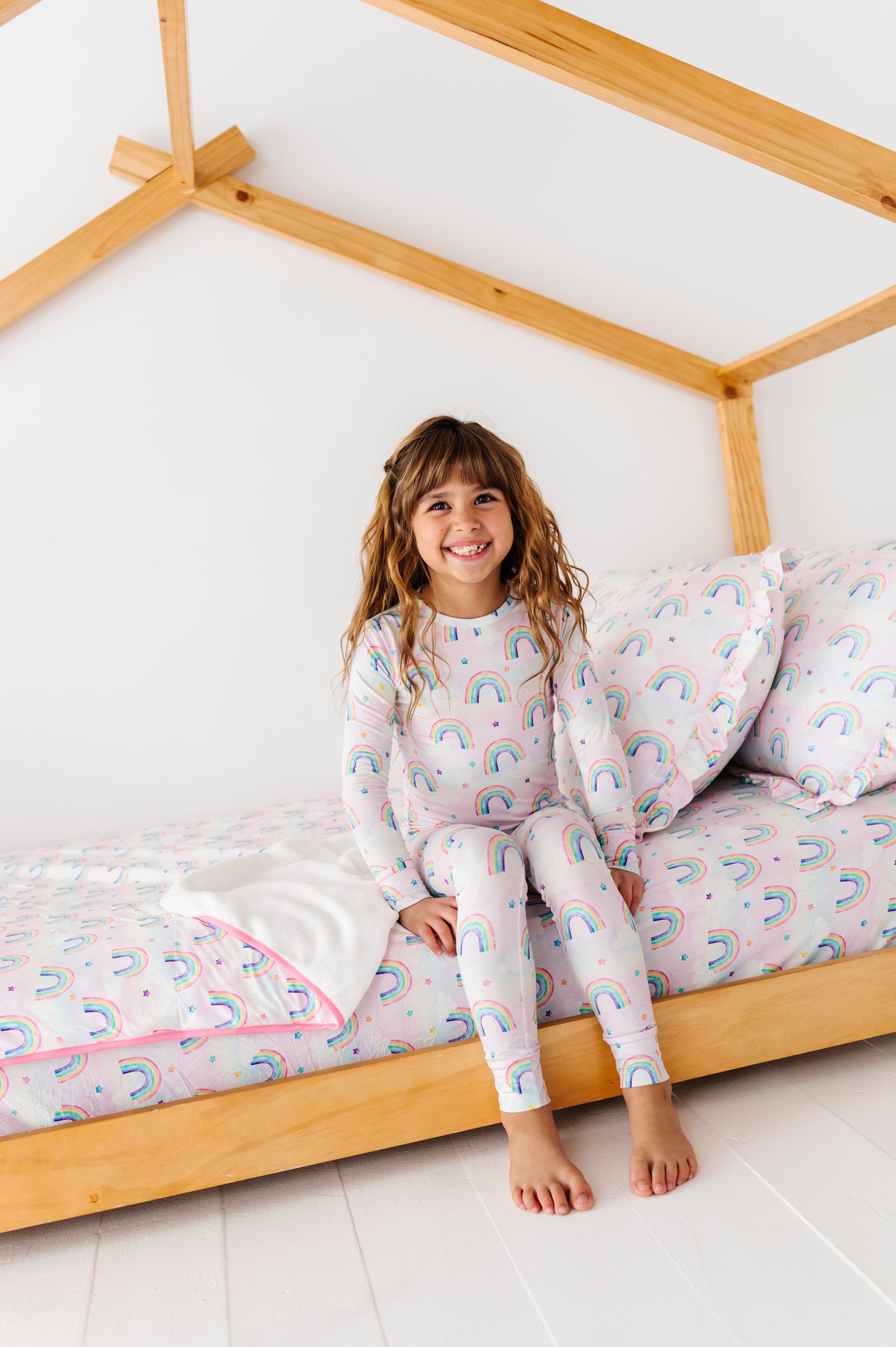 Child in rainbow-themed bamboo pajamas sitting on a bed with wooden frame and matching bamboo bedding.