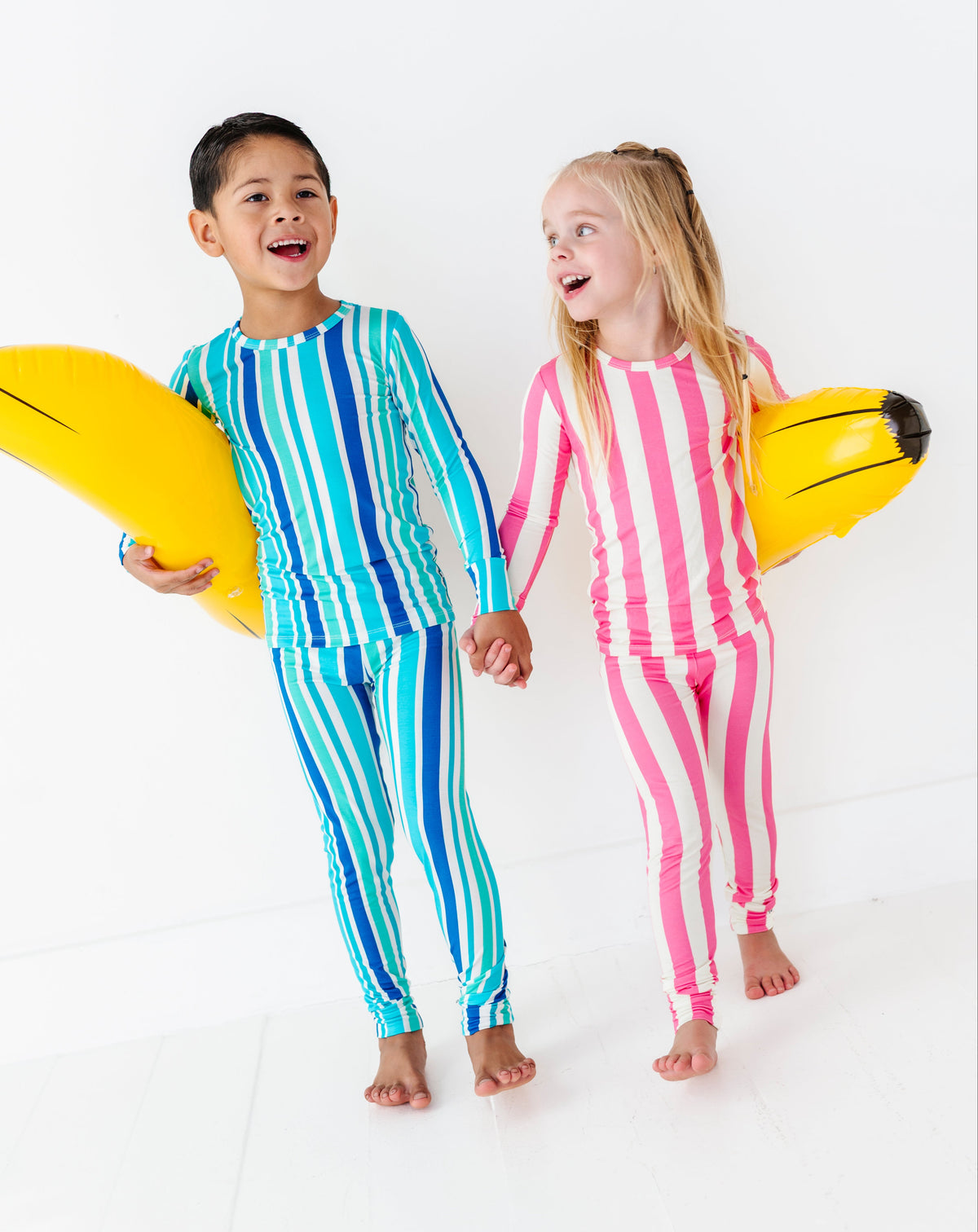 Two children standing barefoot on a white studio floor, each holding a yellow surfboard. The child on the left wears a blue and green vertically striped long-sleeve swim shirt and matching swim pants.