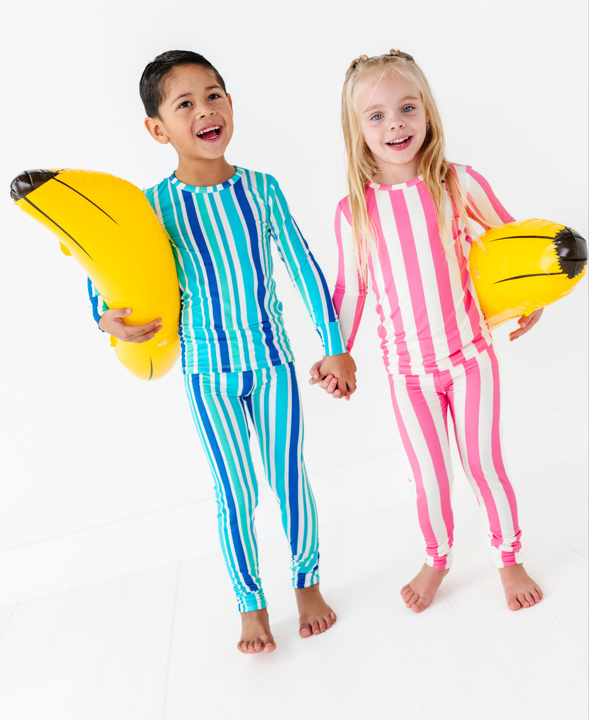 Two children standing on a white studio background, each holding a yellow banana-shaped float. The boy wears a blue and green vertically striped long-sleeve pajama set, and the girl wears a pink and white vertically striped long-sleeve pajama set.