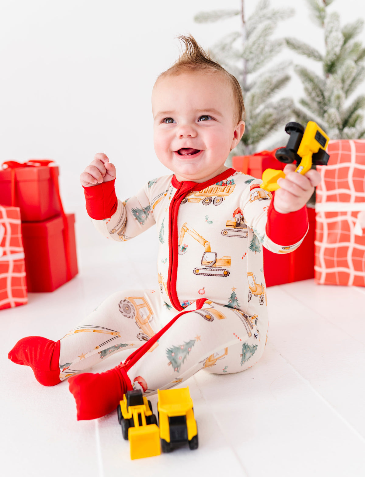 A baby boy wearing a construction-themed bamboo zip romper and playing with construction trucks. He's sitting in front of a Christmas tree and a pile of red wrapped presents 