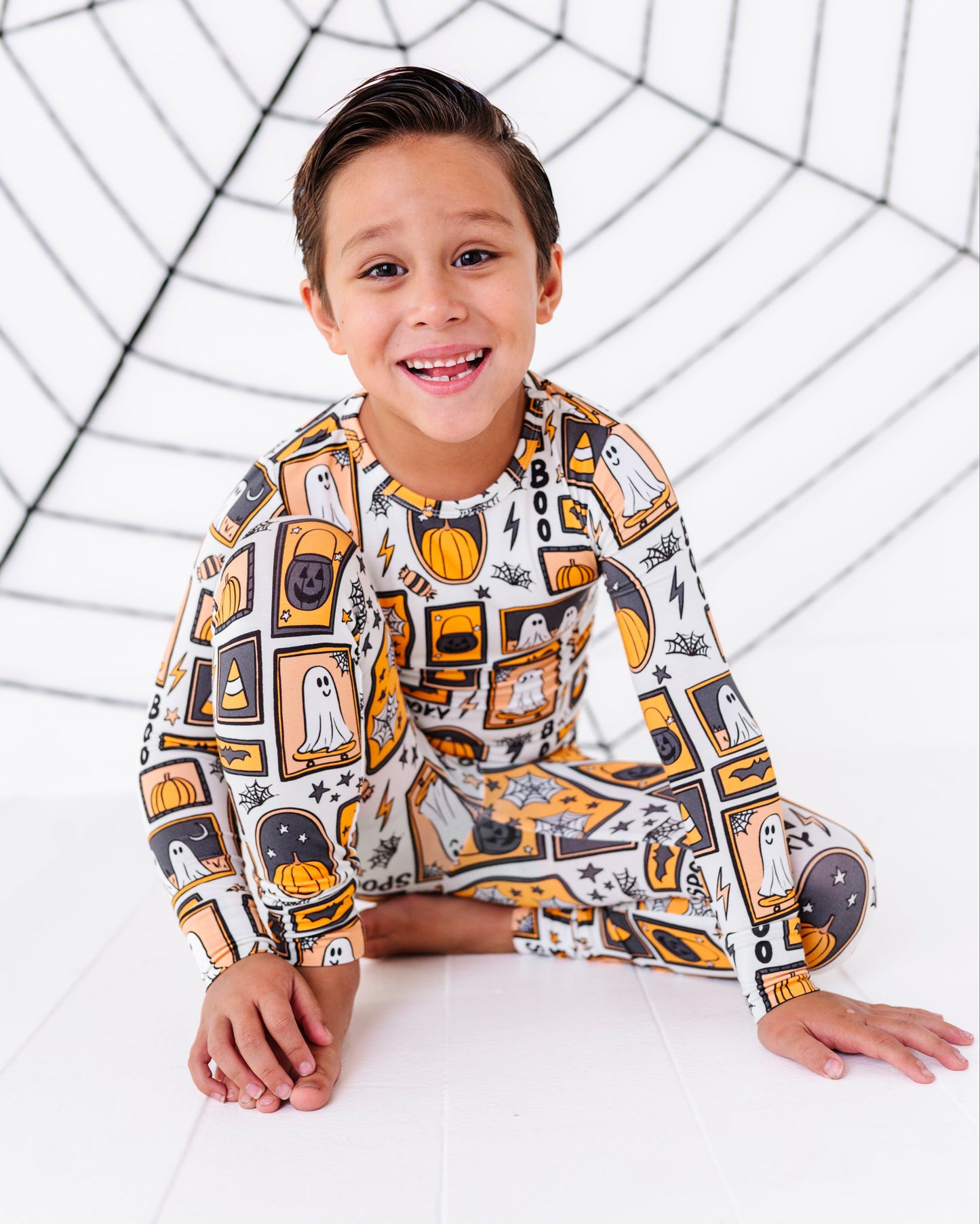 A young boy smiling and sitting on a white studio floor, wearing a long-sleeve pajama set with a Halloween-themed print of ghosts, pumpkins, and spiderwebs. A large black spiderweb graphic is printed on the white background behind him.