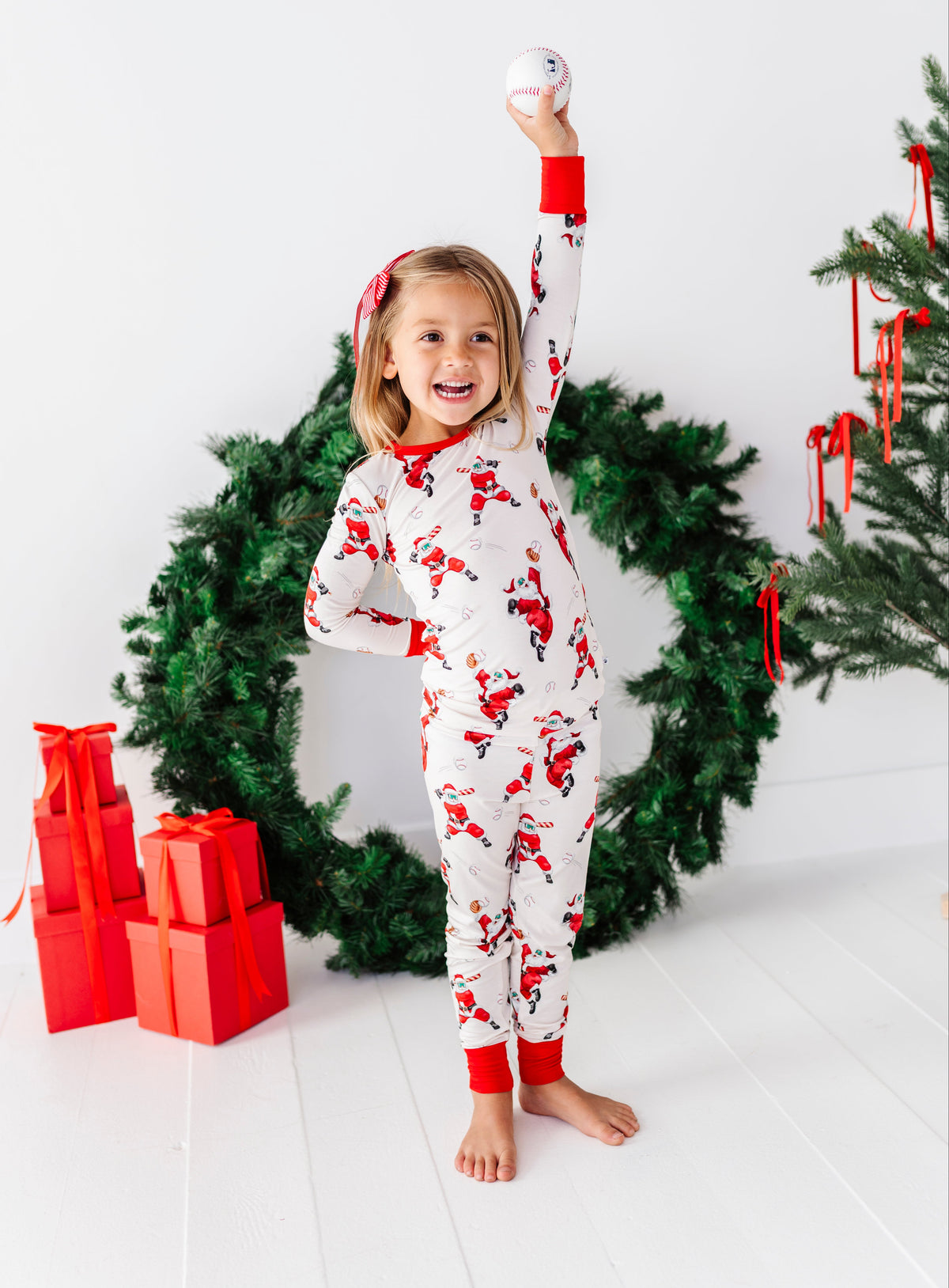 A young girl stands barefoot in a holiday-themed room, wearing a white Santa Claus print one-piece pajama and holding up a white ornament. A green Christmas wreath, a decorated Christmas tree, and red wrapped gift boxes are visible in the background.