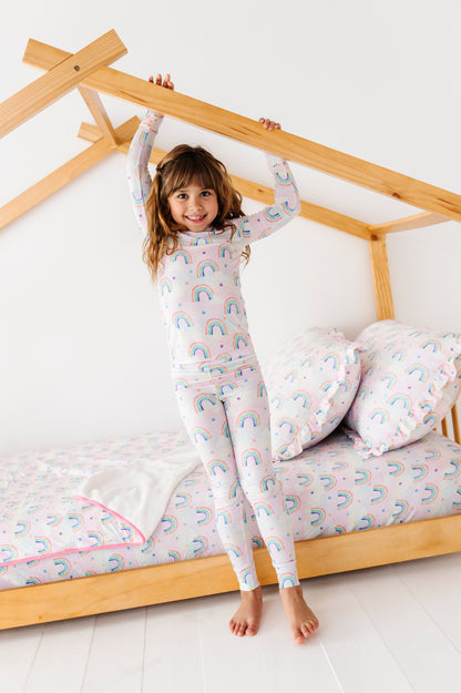 Child in a rainbow-patterned bamboo two-piece pajamas standing on a bed with wooden frame and white walls.