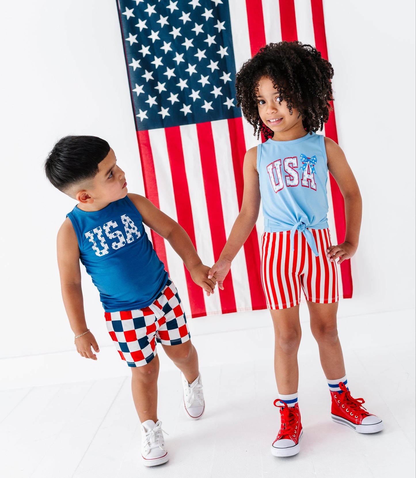 Two children holding hands in front of an American flag, wearing matching 'USA' outfits.