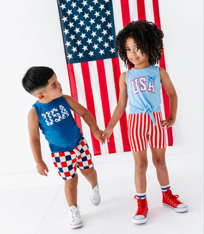 Two children holding hands in front of an American flag, wearing matching 'USA' outfits.