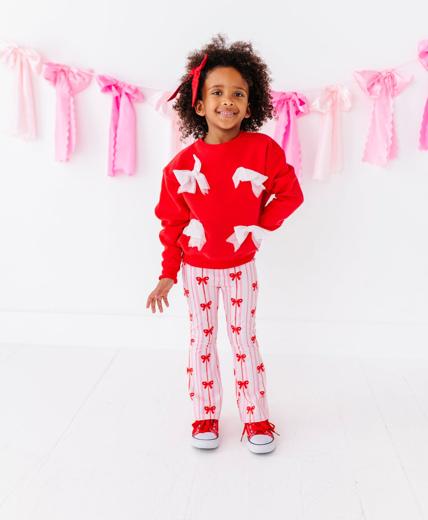 Child wearing a red sweater with white bows and red and white striped bamboo pants standing in front of pink ribbon decorations.