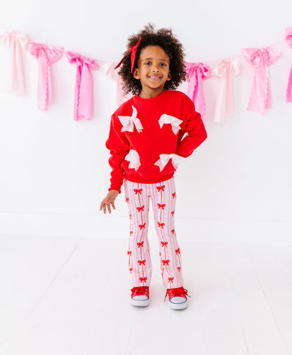 Child wearing a red sweater with white bows and red and white striped bamboo pants standing in front of pink ribbon decorations.