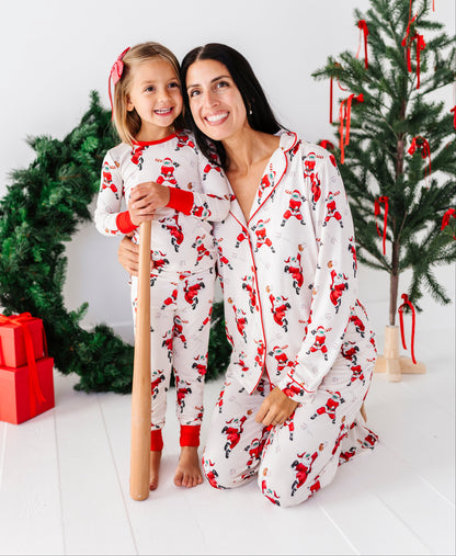 A woman and a young girl wearing matching white Christmas pajamas with a red Santa Claus print, kneeling in a festive holiday room with Christmas trees and wrapped gifts.