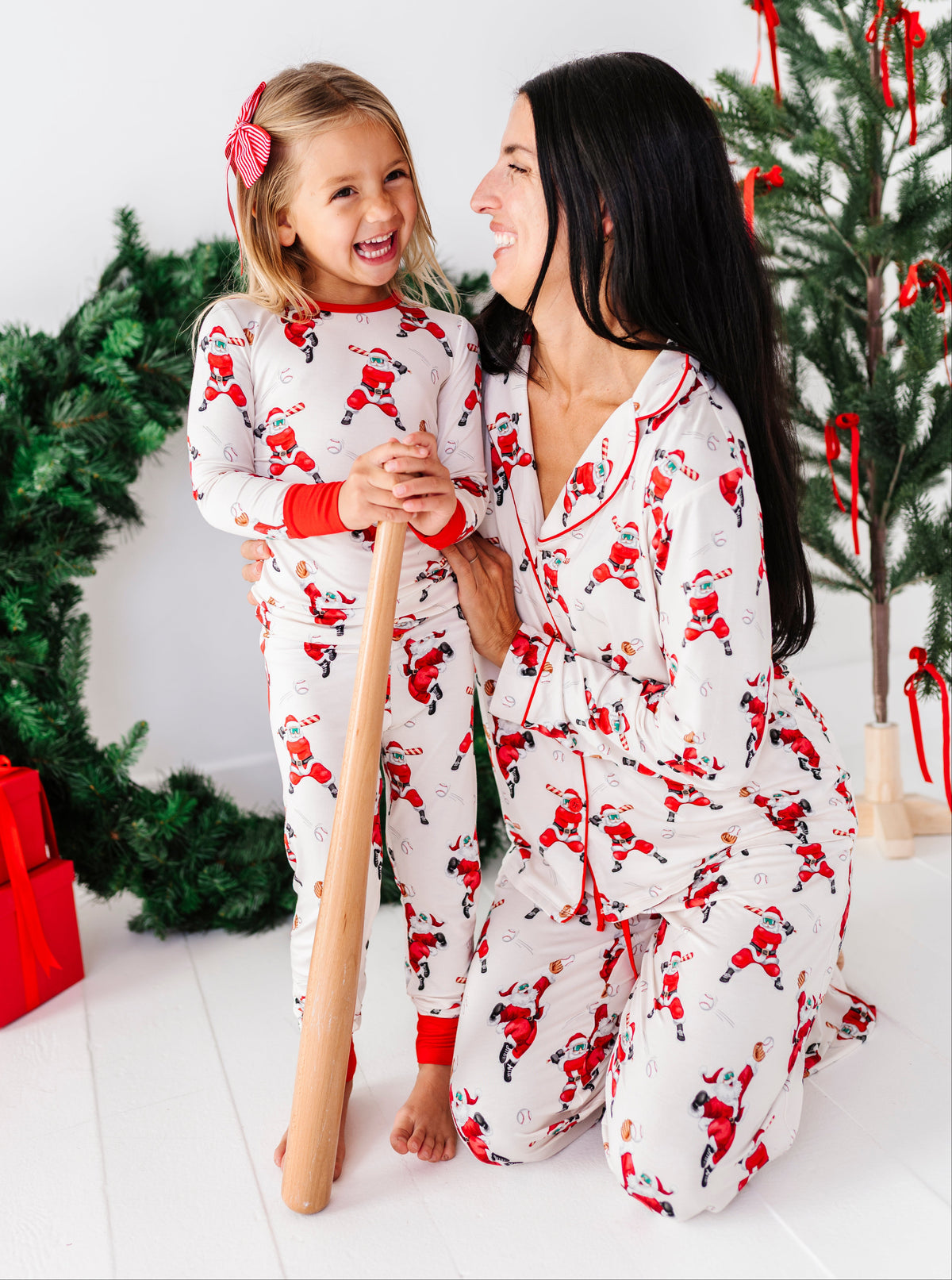 A woman and a young girl wearing matching white Christmas-themed pajamas with a red and black Santa Claus print, posing together in a brightly lit room with a decorated Christmas tree and wreath.