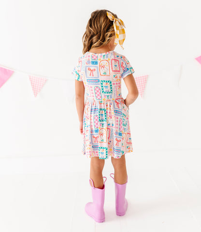 Child wearing a colorful dress with a pattern of various shapes and colors, standing against a white background.