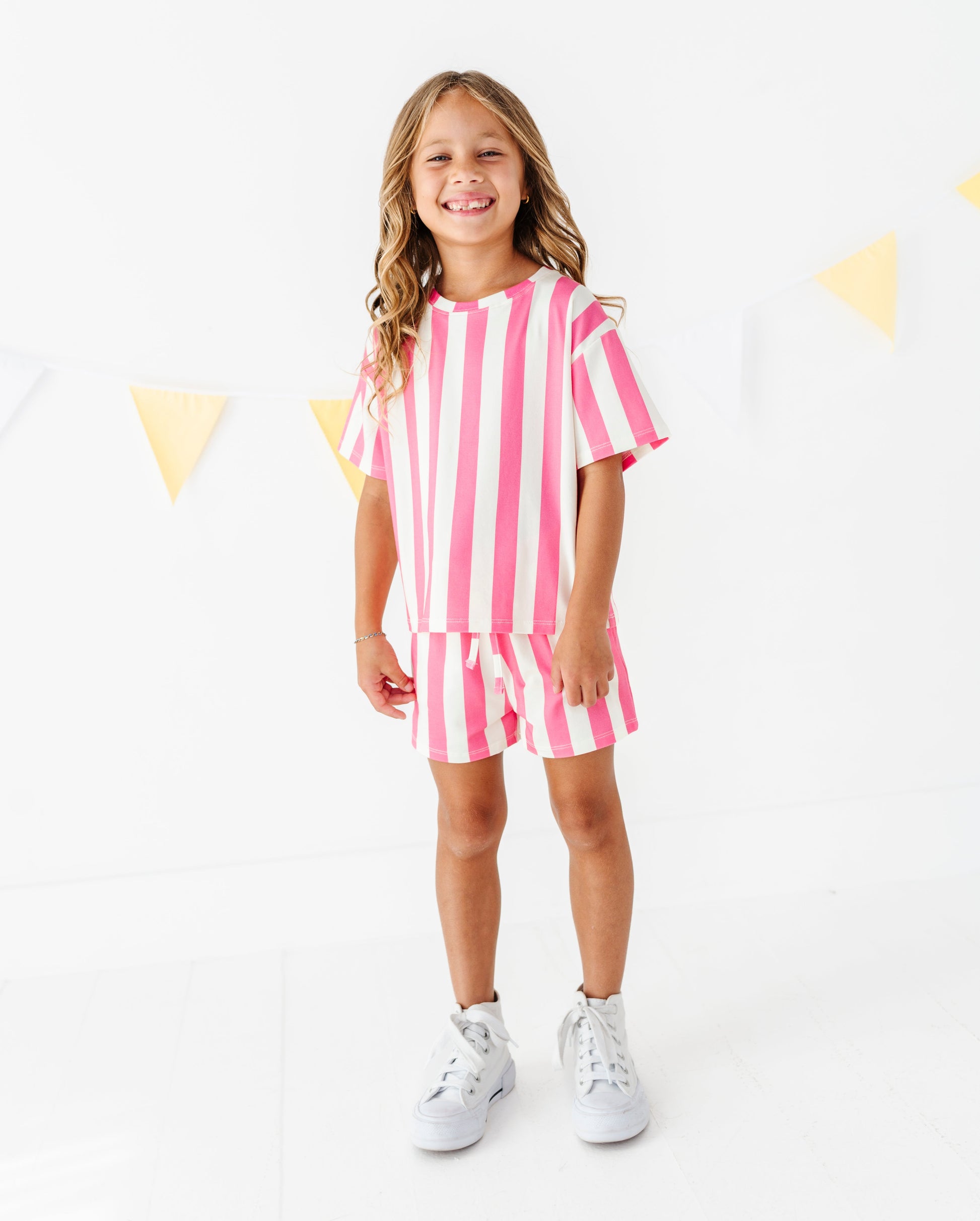 A young girl stands smiling in a bright white studio setting, wearing a matching pink and white vertically striped short-sleeve top and shorts set, paired with white sneakers.