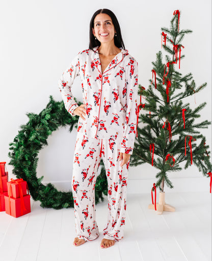 A woman stands in a bright white room wearing a white pajama set with a red Santa Claus print. She is posed between a green Christmas wreath with red wrapped gifts on the left and a decorated Christmas tree on the right.