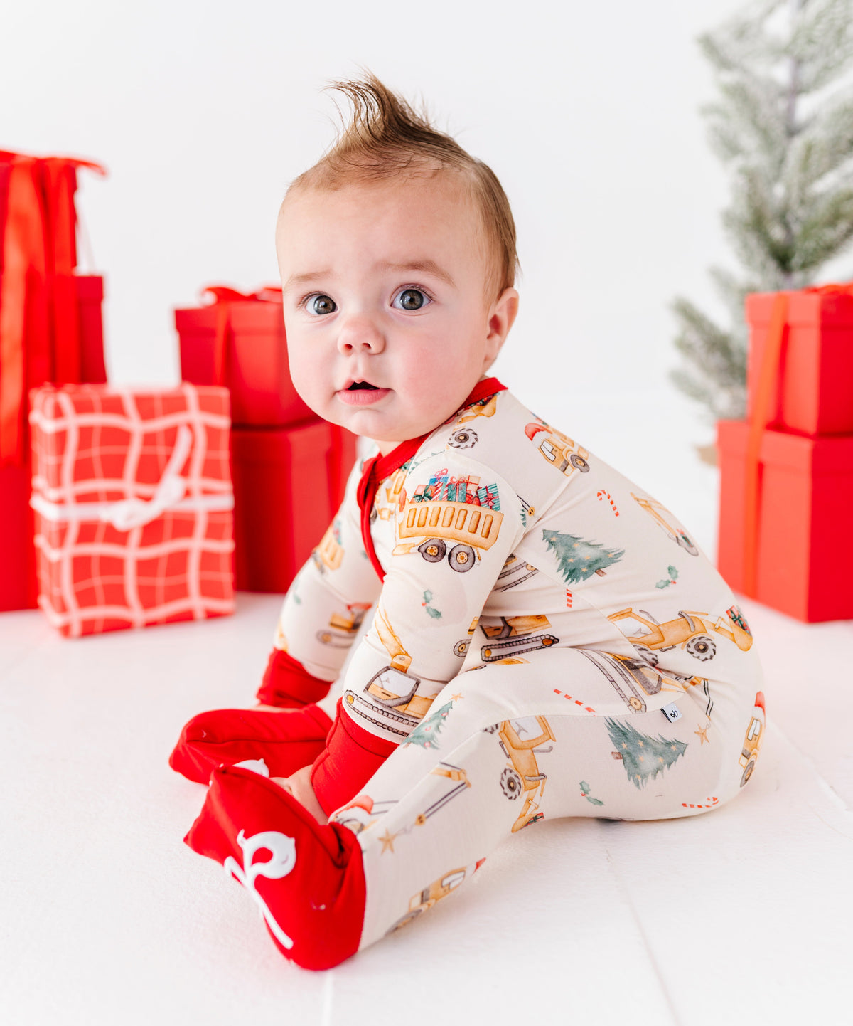 A baby wearing a Christmas construction-themed bamboo convertible romper. He's surrounded by red-wrapped presents and sitting in front of a Christmas tree