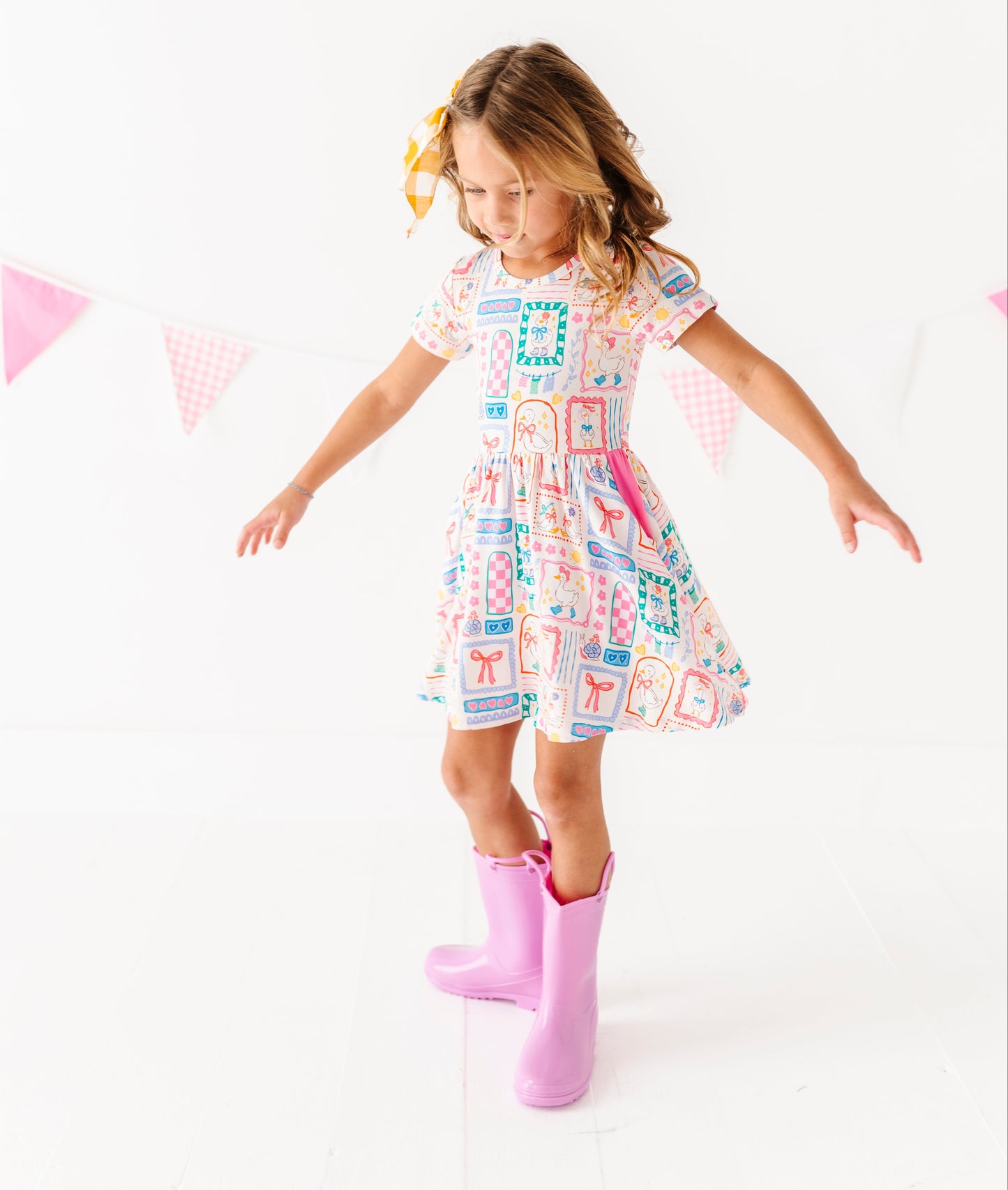 Young girl in a patterned bamboo dress and pink rain boots standing on a white background with small flags.