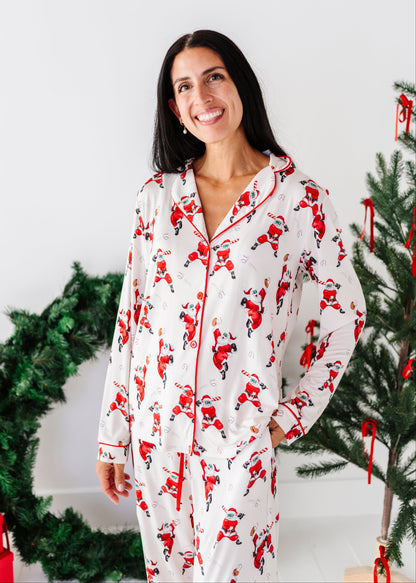 A woman wearing a white long-sleeve pajama set with a red and black Santa Claus print, standing in a holiday-themed setting with a Christmas tree and wreath.