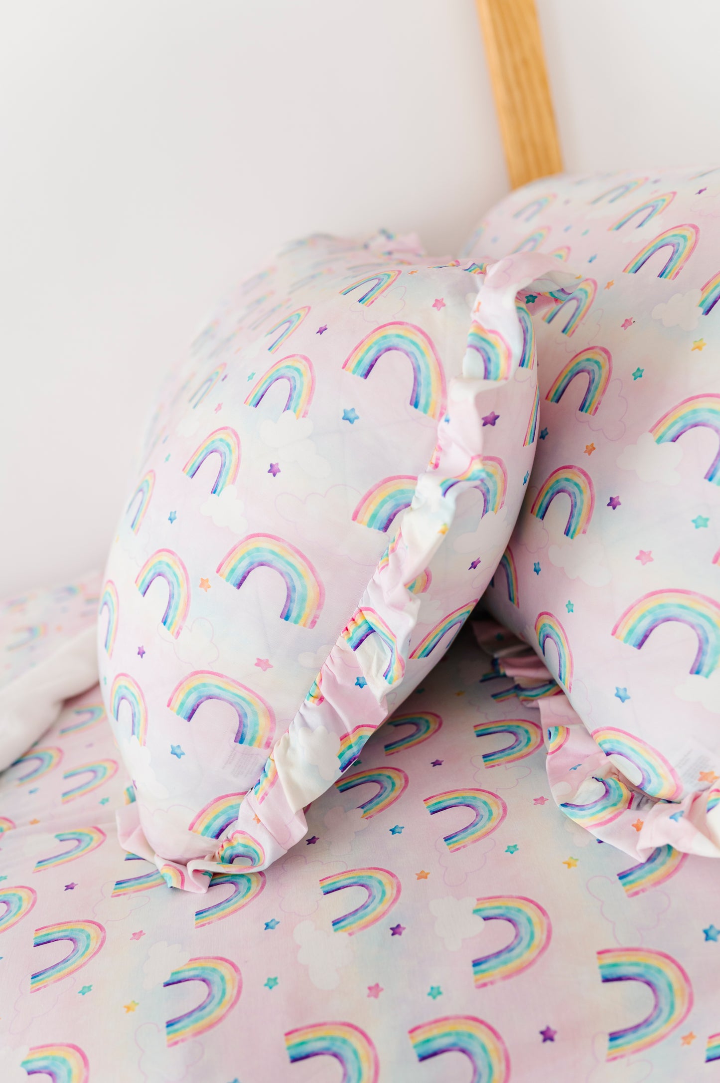 Two Rainbow Skies ruffle zipper pillowcases in a pastel rainbow and star print, styled on a bed with matching bamboo sheets.