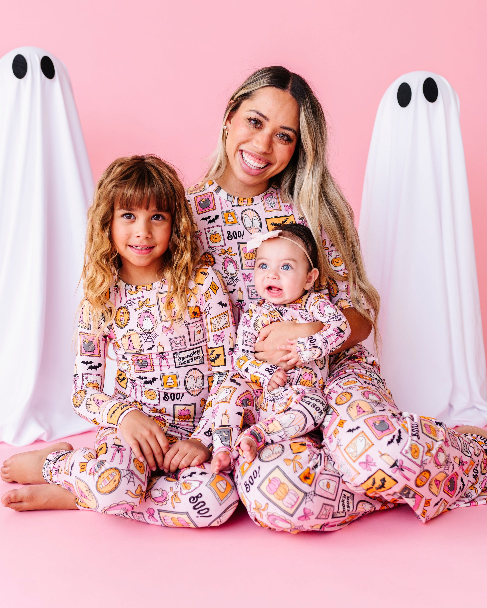 A woman and two children wearing matching pink patterned pajamas sit together on a pink studio floor, flanked by two white ghost props with black eyes. The pajamas have a colorful food and snack collage print.