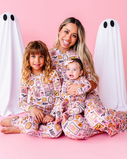 A woman and two children wearing matching pink patterned pajamas sit together on a pink studio floor, flanked by two white ghost props with black eyes. The pajamas have a colorful food and snack collage print.