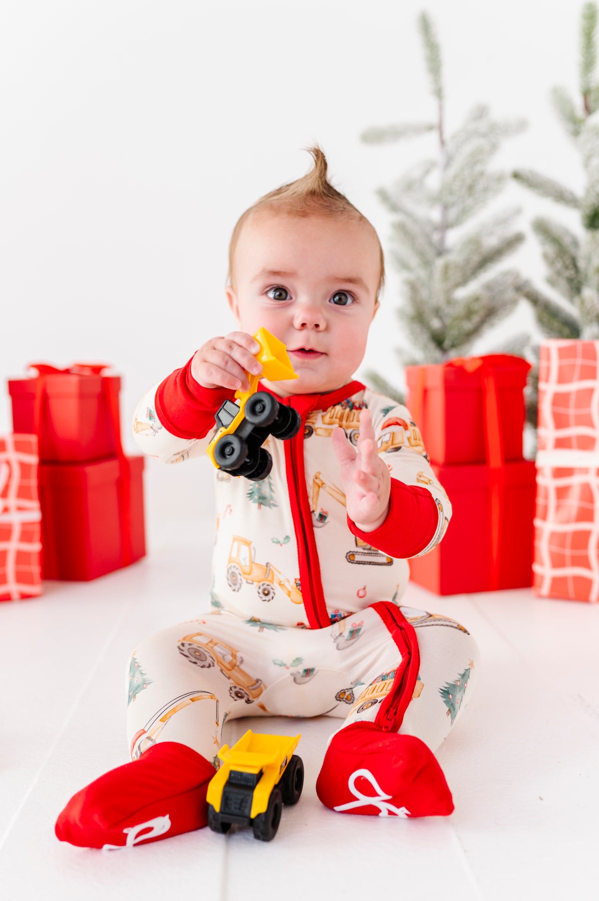 A baby playing with construction trucks while wearing a Christmas construction-themed convertible romper. It's a super soft bamboo zip romper 