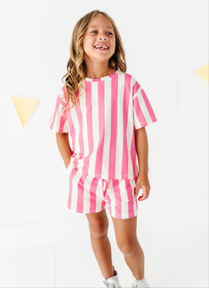 A young girl smiling and looking to the side, wearing a matching pink and white vertically striped short-sleeve t-shirt and shorts set, standing against a white wall with subtle yellow bunting.