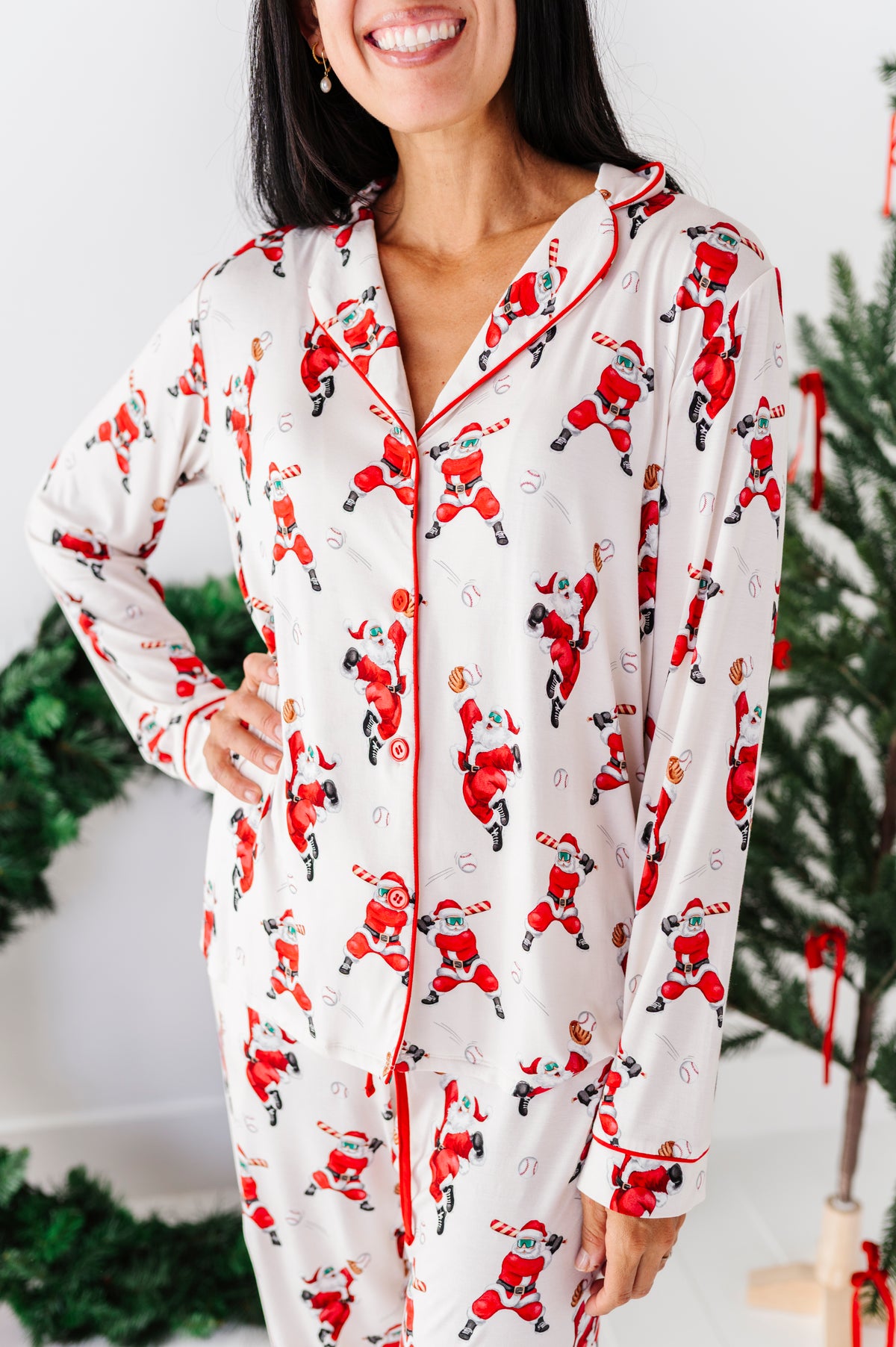 A woman wearing a long-sleeve white pajama set with a red and black Santa Claus print, standing in front of a decorated Christmas tree.