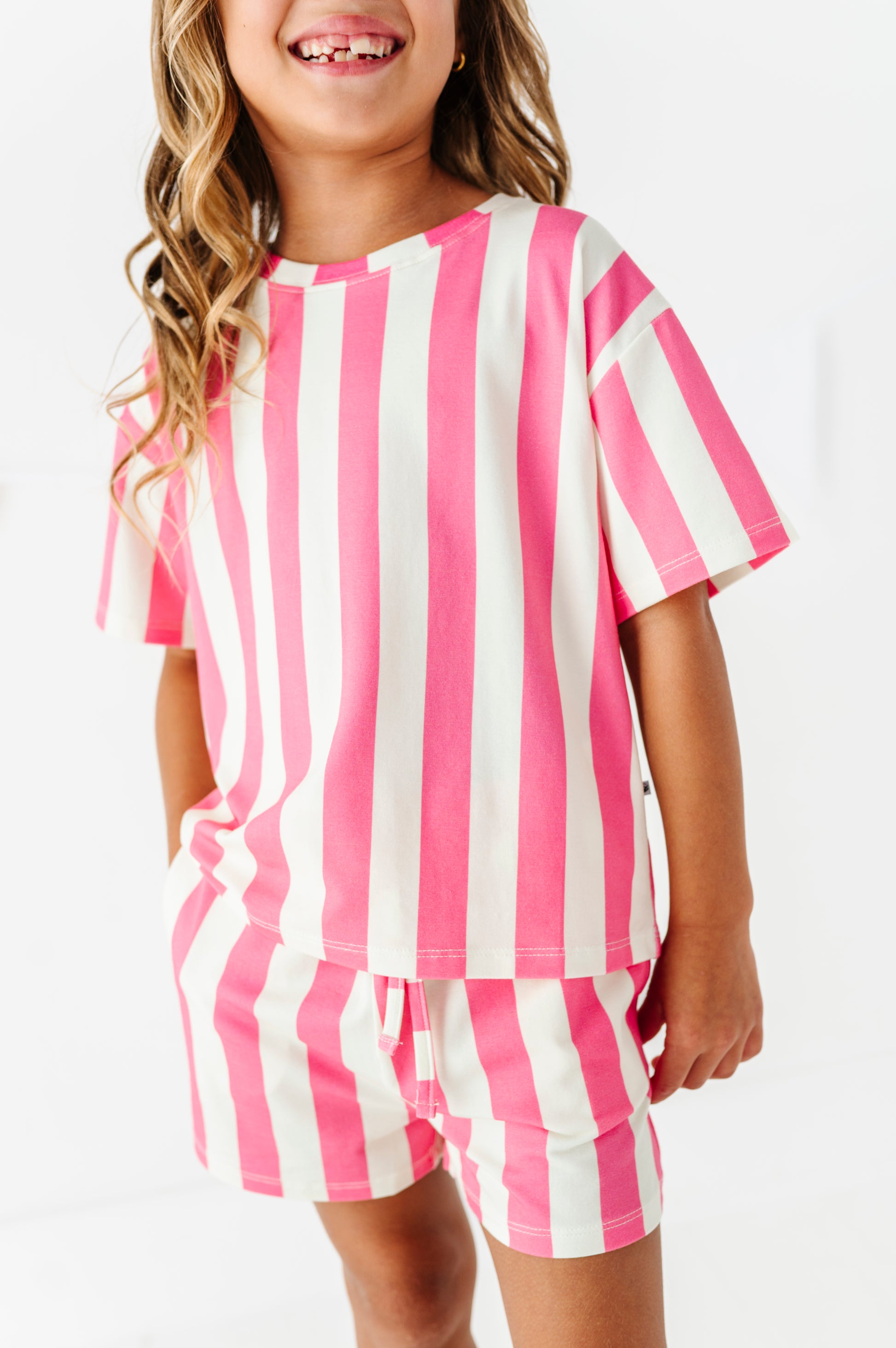 A young girl wearing a matching two-piece pajama set with pink and white vertical stripes, featuring a short-sleeve top and shorts, against a plain white background.