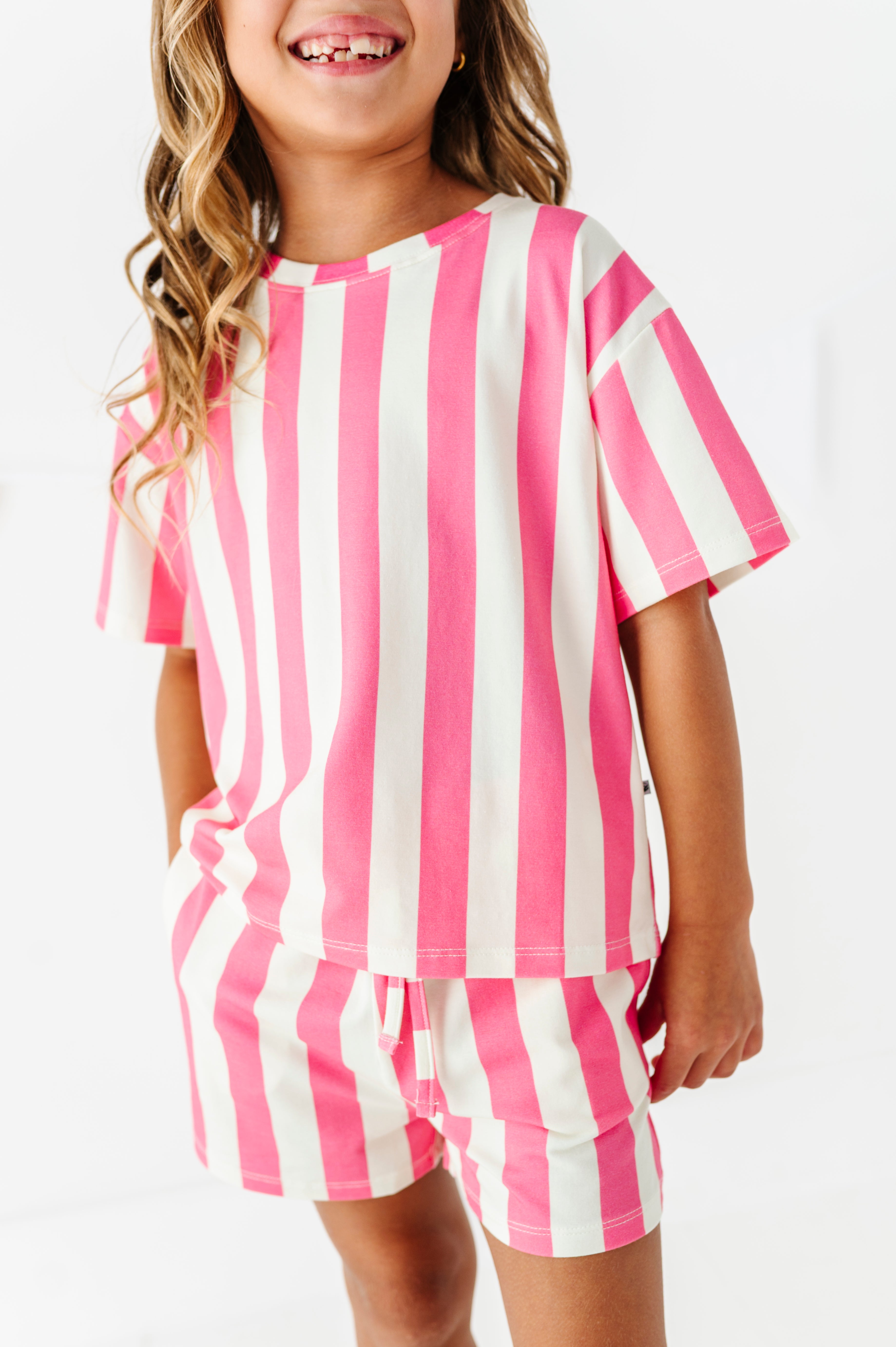 A young girl wearing a matching two-piece pajama set with pink and white vertical stripes, featuring a short-sleeve top and shorts, against a plain white background.