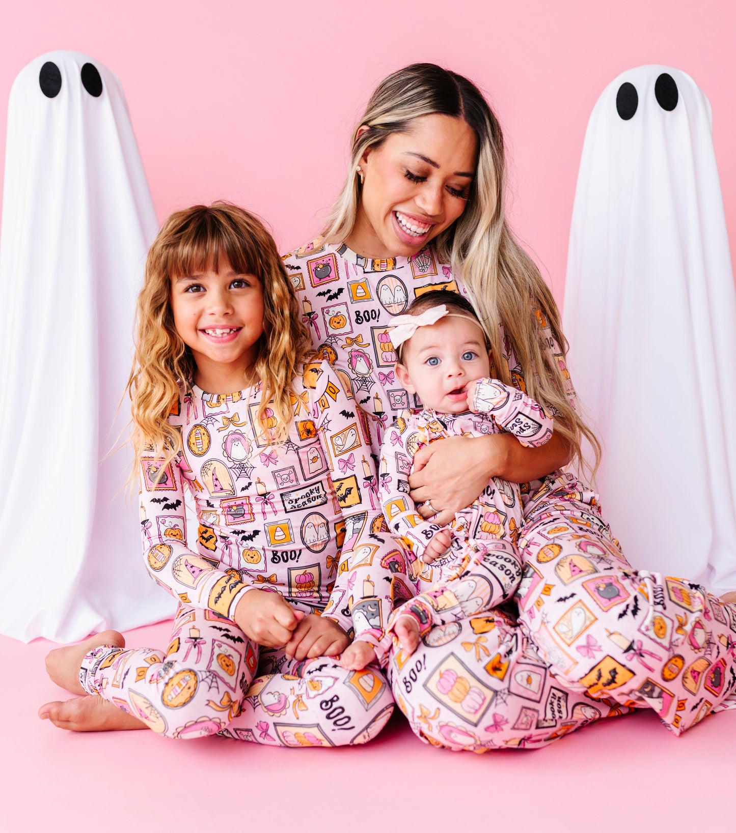 A woman and two children wearing matching pink patterned pajamas sit on a pink floor in front of a pink backdrop with white ghost props. The pajama set has a colorful cartoon-style print with text and illustrations.