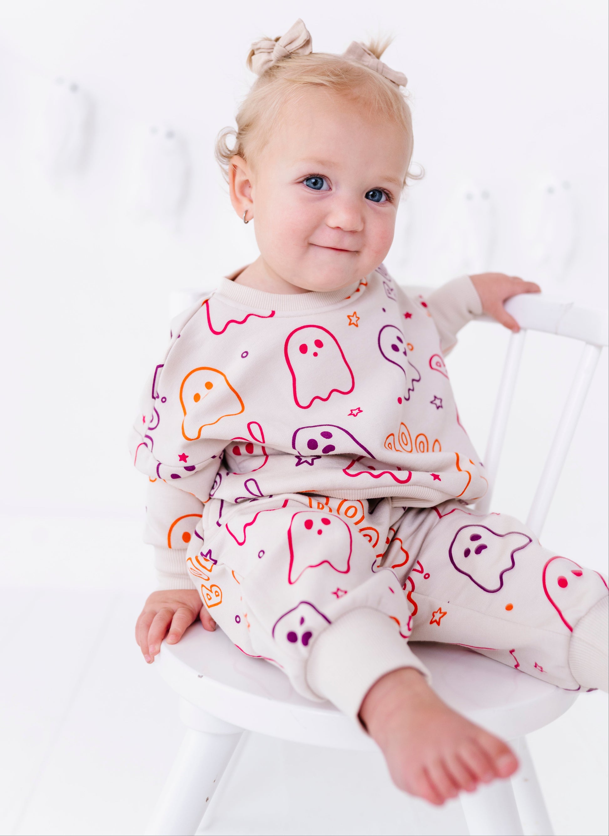 A toddler girl with blonde pigtails sits on a white wooden chair, wearing a matching cream-colored pajama set with a colorful ghost pattern.