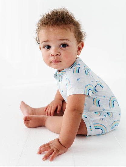 Baby sitting on a white surface wearing a light blue bamboo romper with rainbow patterns.