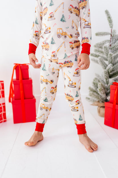 A child wearing a long-sleeve and matching pants bamboo pajama set with a holiday construction theme, featuring yellow construction vehicles and trees on an off-white background with red cuffs and ankles, standing in a festive room with wrapped gifts and a Christmas tree.