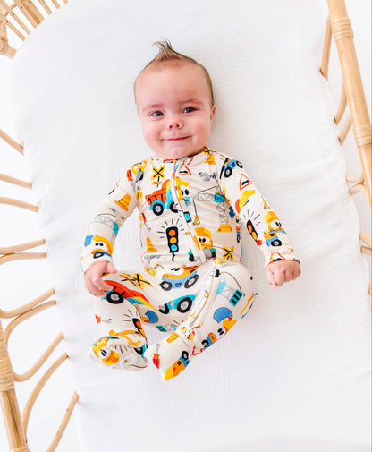 A baby lying on a white cushion inside a rattan bassinet, wearing a cream-colored footed sleeper with a colorful vehicle print.
