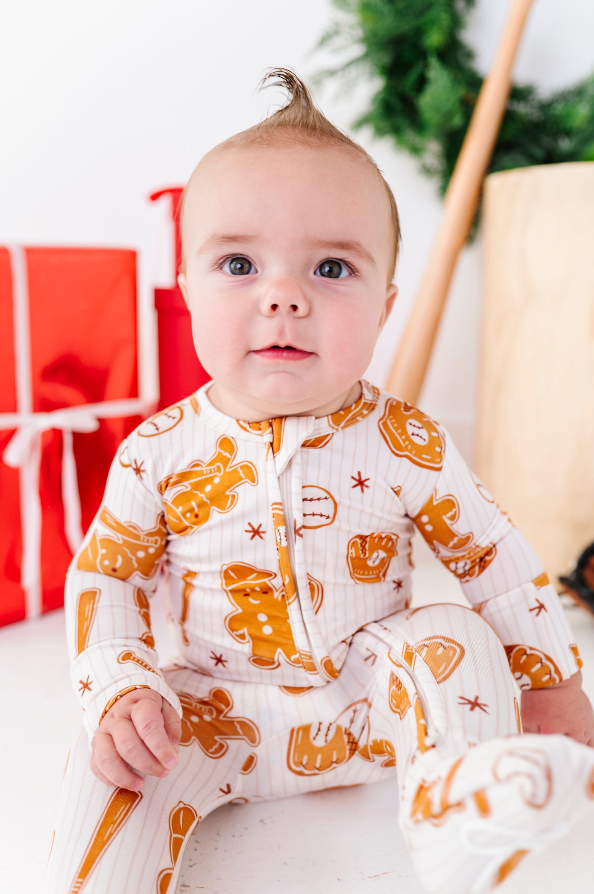 A baby wearing a white long-sleeve footless onesie with an orange gingerbread cookie print, sitting in a festive indoor setting with wrapped gifts and holiday decor in the background.