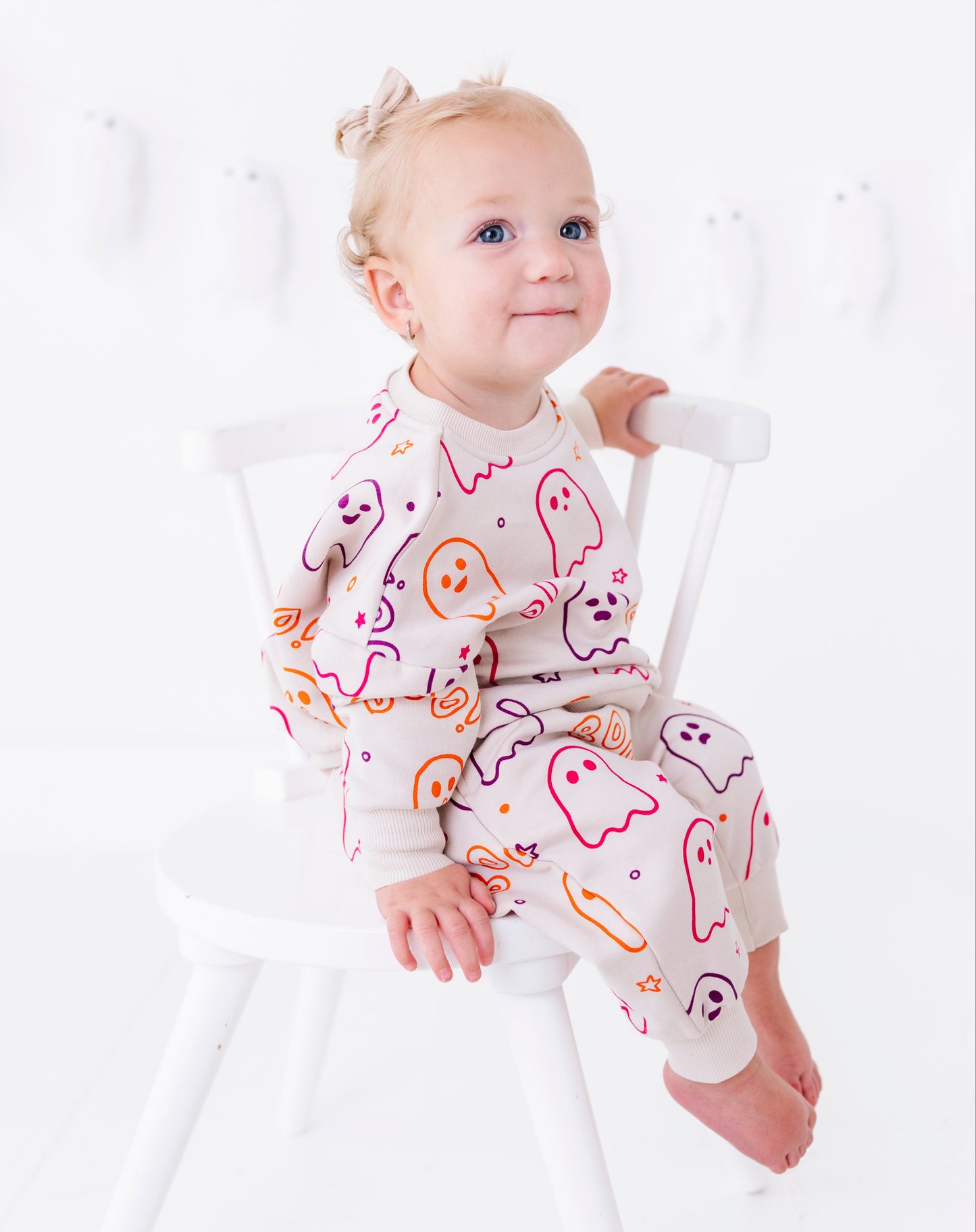 A baby girl sitting on a white wooden chair, wearing a light beige long-sleeve pajama set with a colorful ghost and pumpkin pattern.