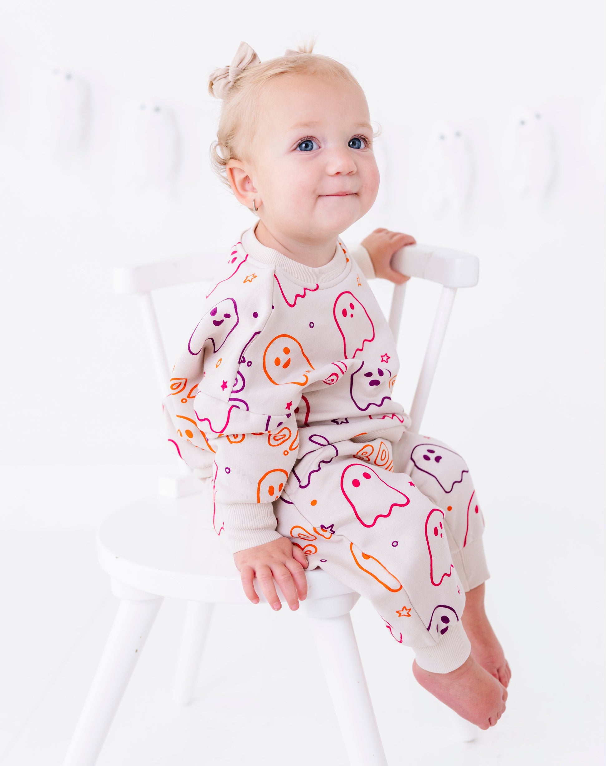 A baby girl sitting on a white wooden chair, wearing a light beige long-sleeve pajama set with a colorful ghost and pumpkin pattern.
